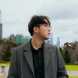 A man wearing a gray coat looks to the right with a park and cityscape behind him. 