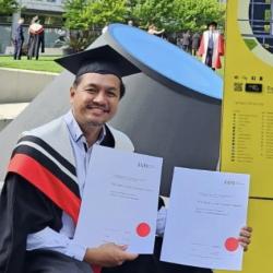 Man in graduation garb holding up two diplomas.