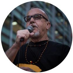 Indigenous man wearing a black tshirt and black glasses, holding and speaking into a microphone.