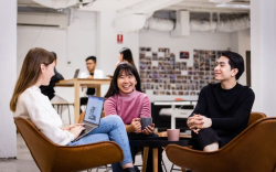 Three students sitting on on chairs in a circle, laughing and smiling. Two students are drinking a cup of coffee and one is working on a laptop.