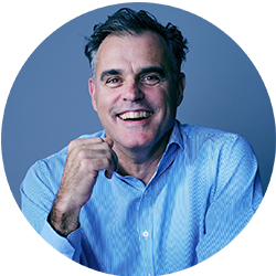 Man in a blue business shirt looking and smiling at the camera with his hand up close to his face