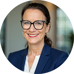 Woman with hair tied back wearing black glasses, white shirt and navy suit jacket, smiling and looking at the camera