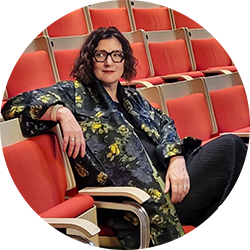 Woman sitting in a theatre looking off camera. She has short brown hair is wearing glasses and a yellow and black flower print shirt with a black skirt. 