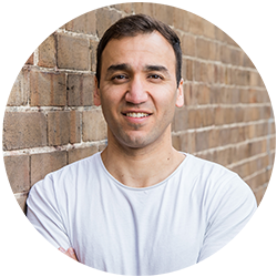 Man in a white tshirt leaning against a wall smiling and looking at the camera