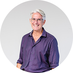 Man with white hair and grey glasses in a purple shirt smiling and looking at the camera