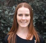 A headshot of Claire, dressed in a simple black jacket smiles widely against a background of green bushes.