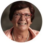 Pat Turner AM smiling. She is wearing a pair of red-framed glasses, a light pink shirt, and a pearl necklace. 