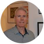 Stephen Peterson has short grey hair and he is wearing a button up shirt. He is standing in front of a gallery wall of paintings.