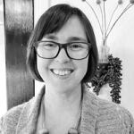 Black and white photo of Naomi Deck smiling at the camera, taken from the shoulders up. Naomi wears glassess with a dark-coloured frame and has a bob haircut. She is wearing a cardigan.