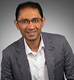 Man smiling and looking at the camera wearing glasses and a pin stripe suit jacket with a white business shirt